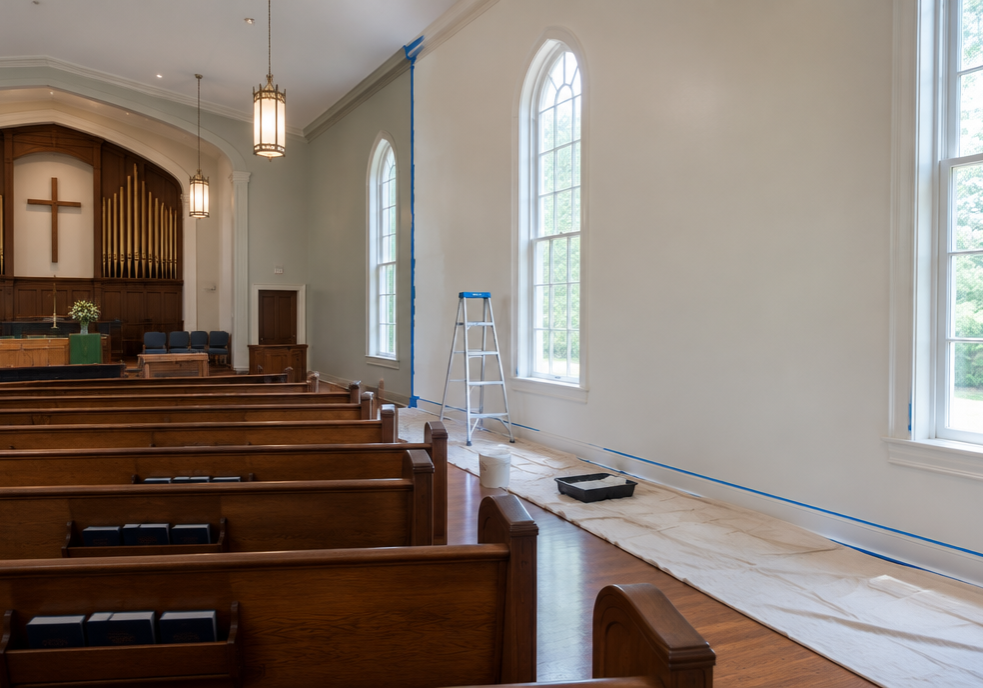 Empty church sanctuary during a phased weekday repaint with one wall freshly painted, drop cloths and a ladder set aside